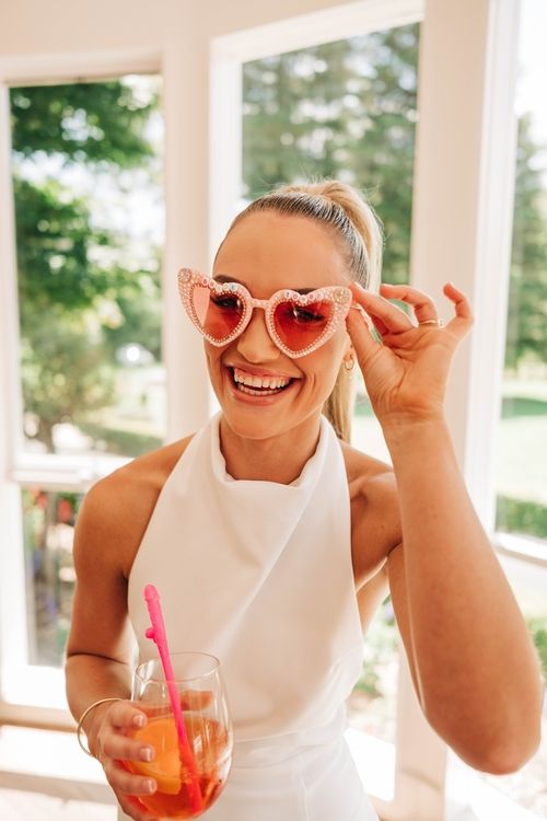 Woman in white outfit wearing heart-shaped sunglasses holding a cocktail.