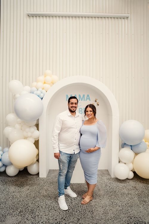 A couple poses in front of a white arch decorated with balloon garlands in grey, white and peach colors.