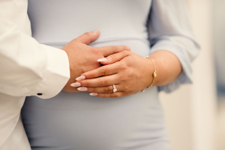 Close-up of hands with wedding rings and a baby bump in grey dress.
