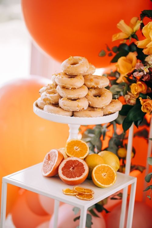 Glazed donuts displayed on a white cake stand with citrus fruits and orange flowers.