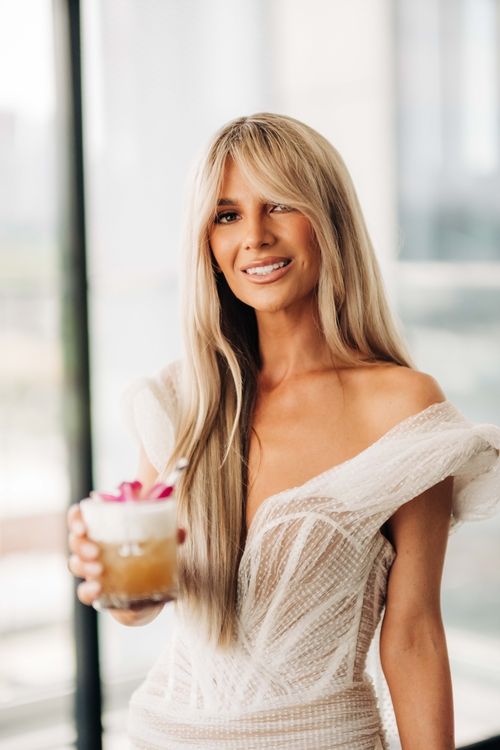 A woman in a white dress holds a cocktail while posing by a window.
