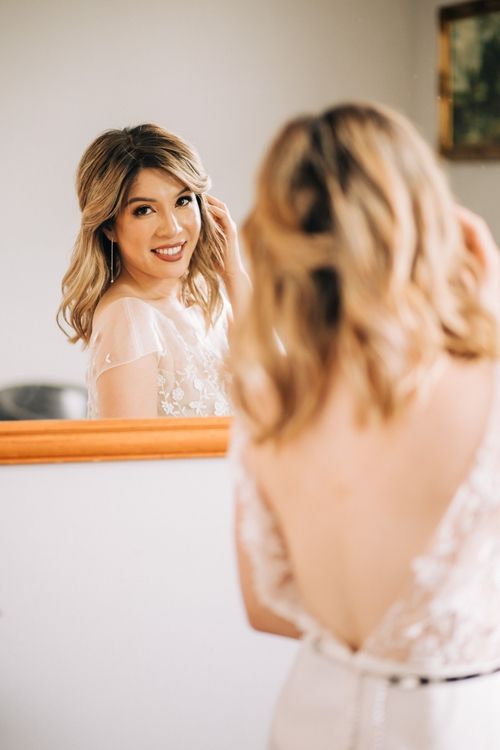 A reflection in a mirror shows a woman in a delicate lace wedding dress with soft makeup and wavy blonde hair.