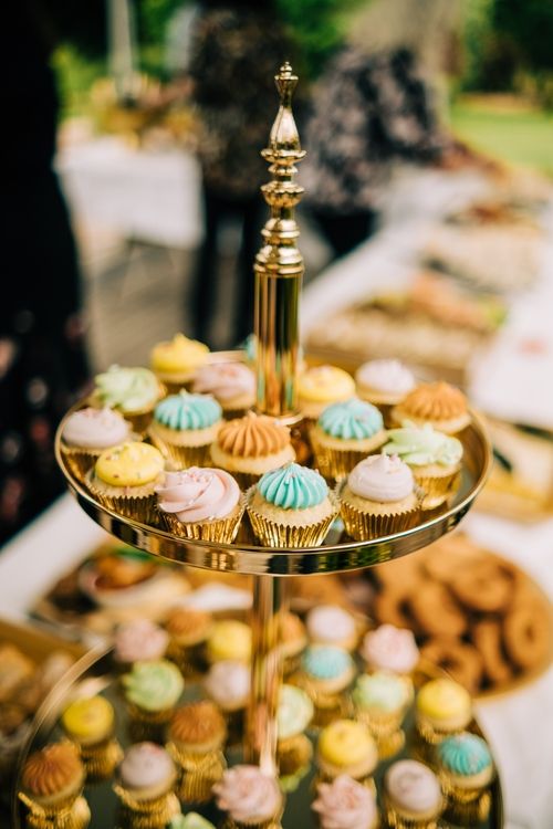Elegant tiered gold dessert stand displaying colorful mini cupcakes at a party reception.