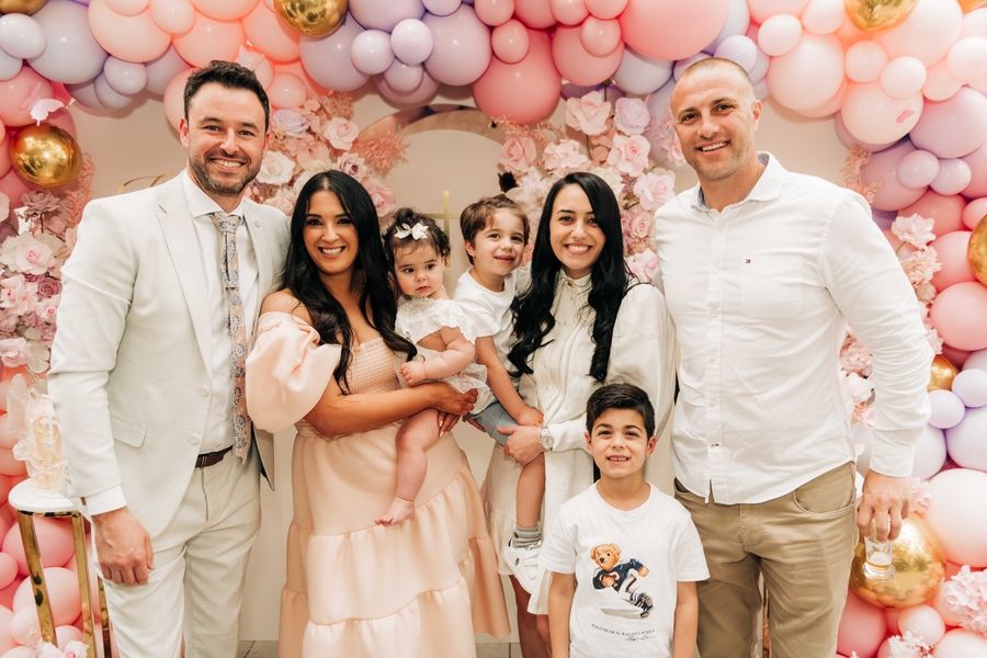 Family gathers for a celebration photo in front of pink balloon arch and floral decorations.