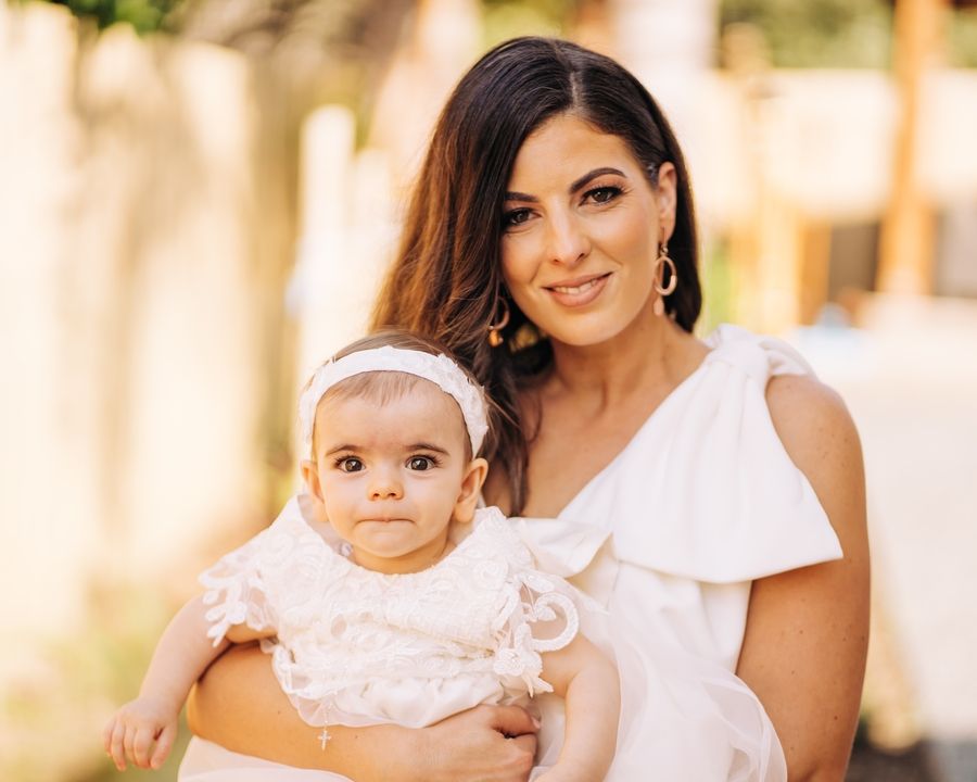 Baby in white dress poses with adult for formal portrait outdoors.