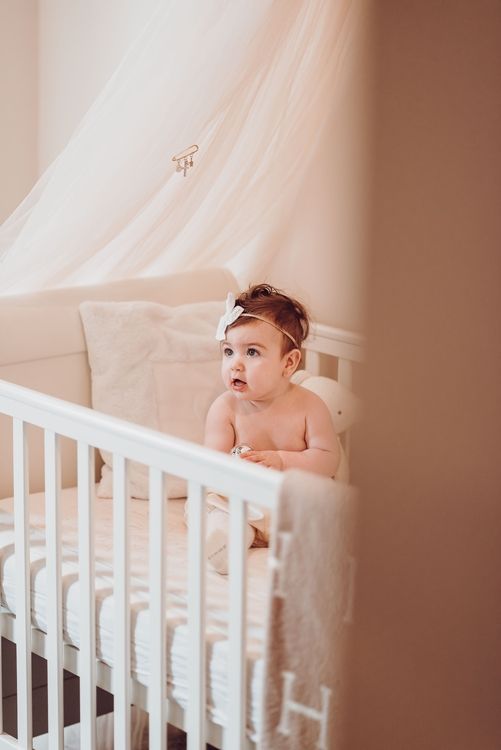 Baby in white headband sits alert in white crib against soft pink wall.