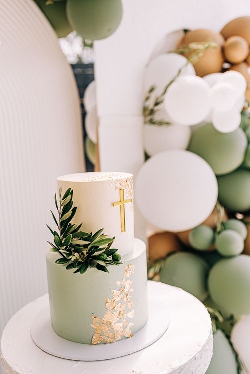 Elegant christening cake decorated with gold cross and green foliage against balloon backdrop.