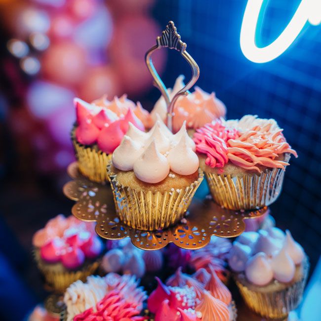Colorful cupcakes with pink frosting displayed on a tiered stand under neon lighting.