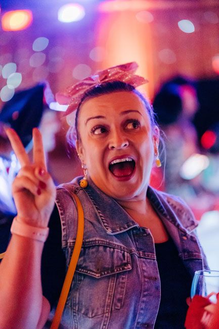Person in denim jacket making peace sign while enjoying a party with colorful lighting.