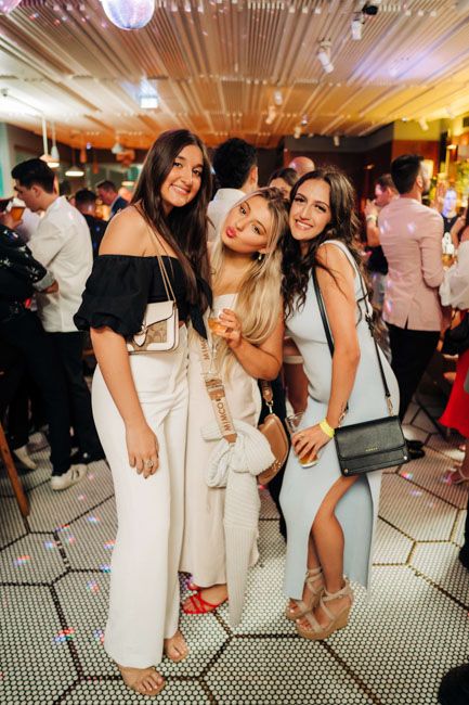 Three friends posing together at a lively party venue with decorative lighting and festive atmosphere.