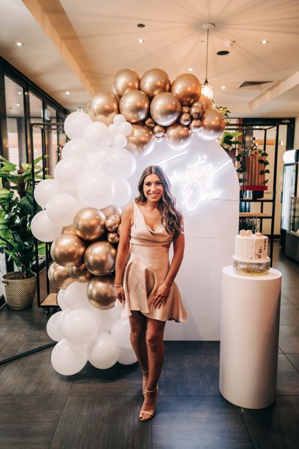 Woman in champagne colored dress poses under elegant balloon arch with cake display.