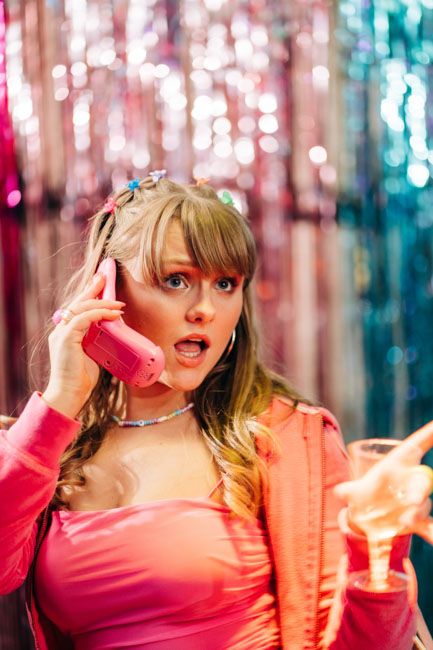Woman in pink outfit makes expressive face while holding phone at party.