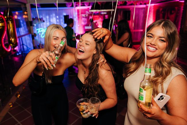 Three friends enjoying drinks and laughter at a vibrant nightclub with pink lighting.
