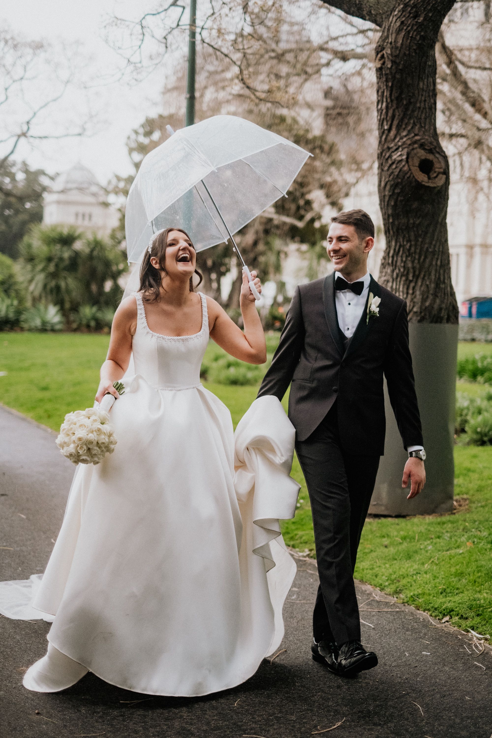 A newly married couple walk and laugh beneath a white umbrella.
