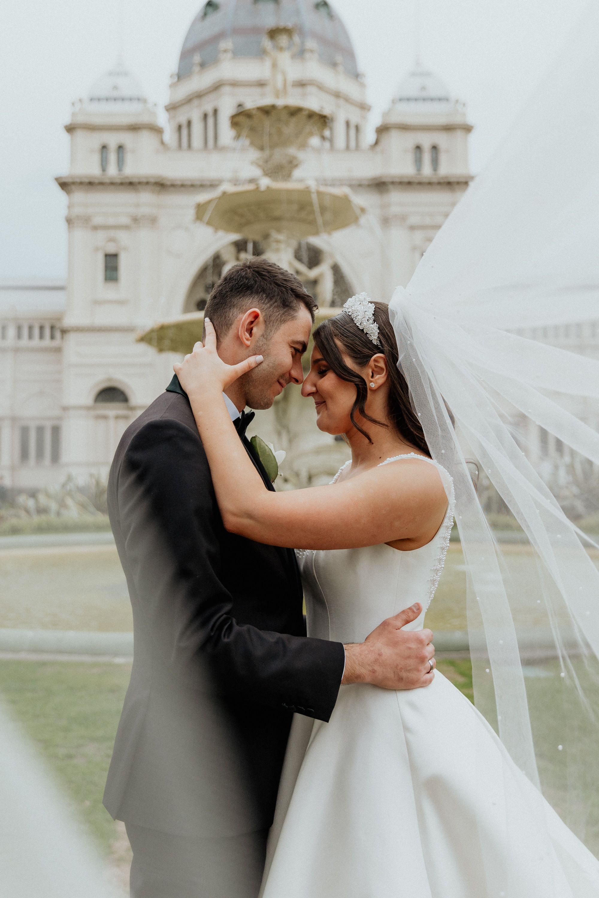 A groom kisses his bride on the forehead beside a fountain.
