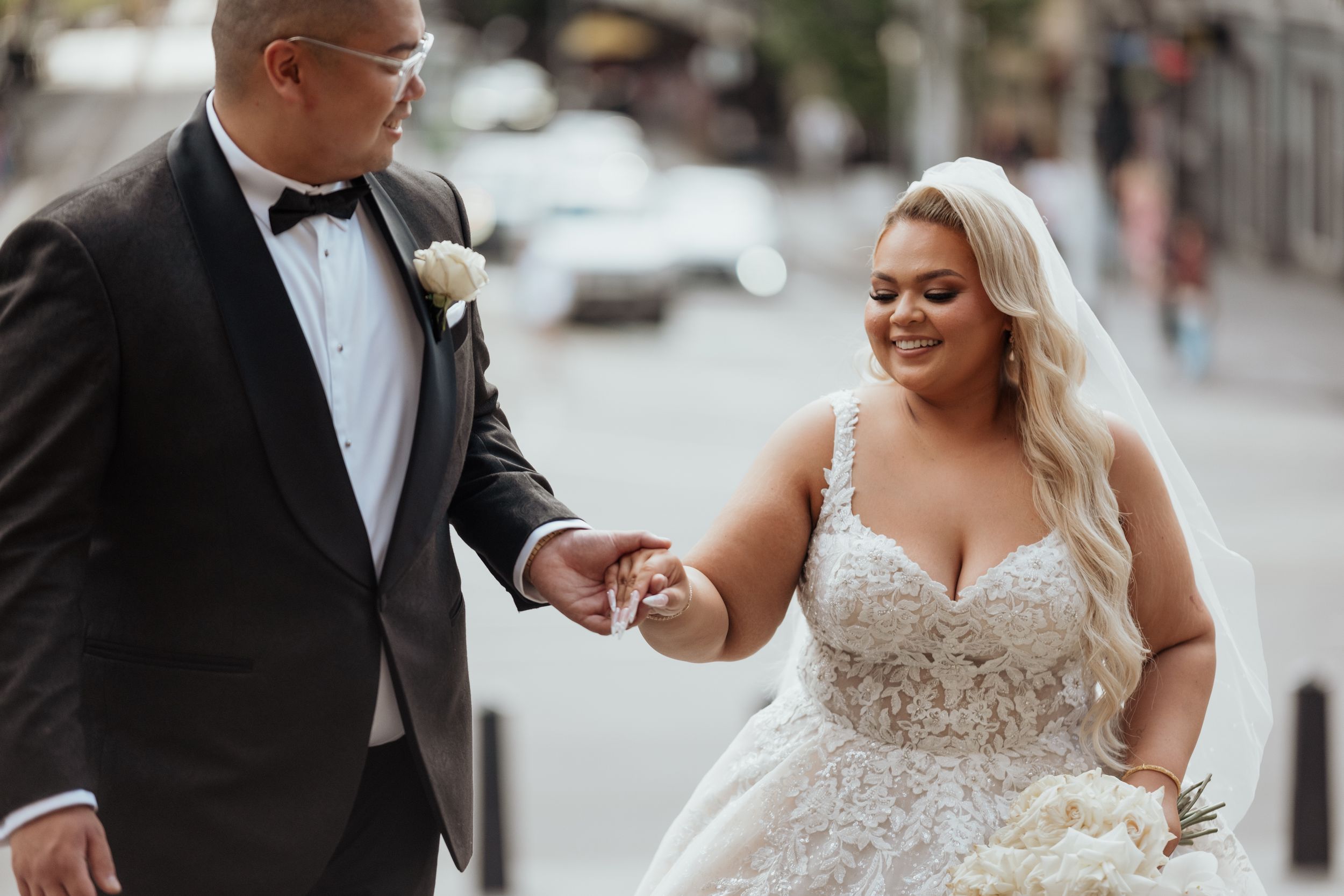 A newly married couple walk hand in hand and smile on a city street.