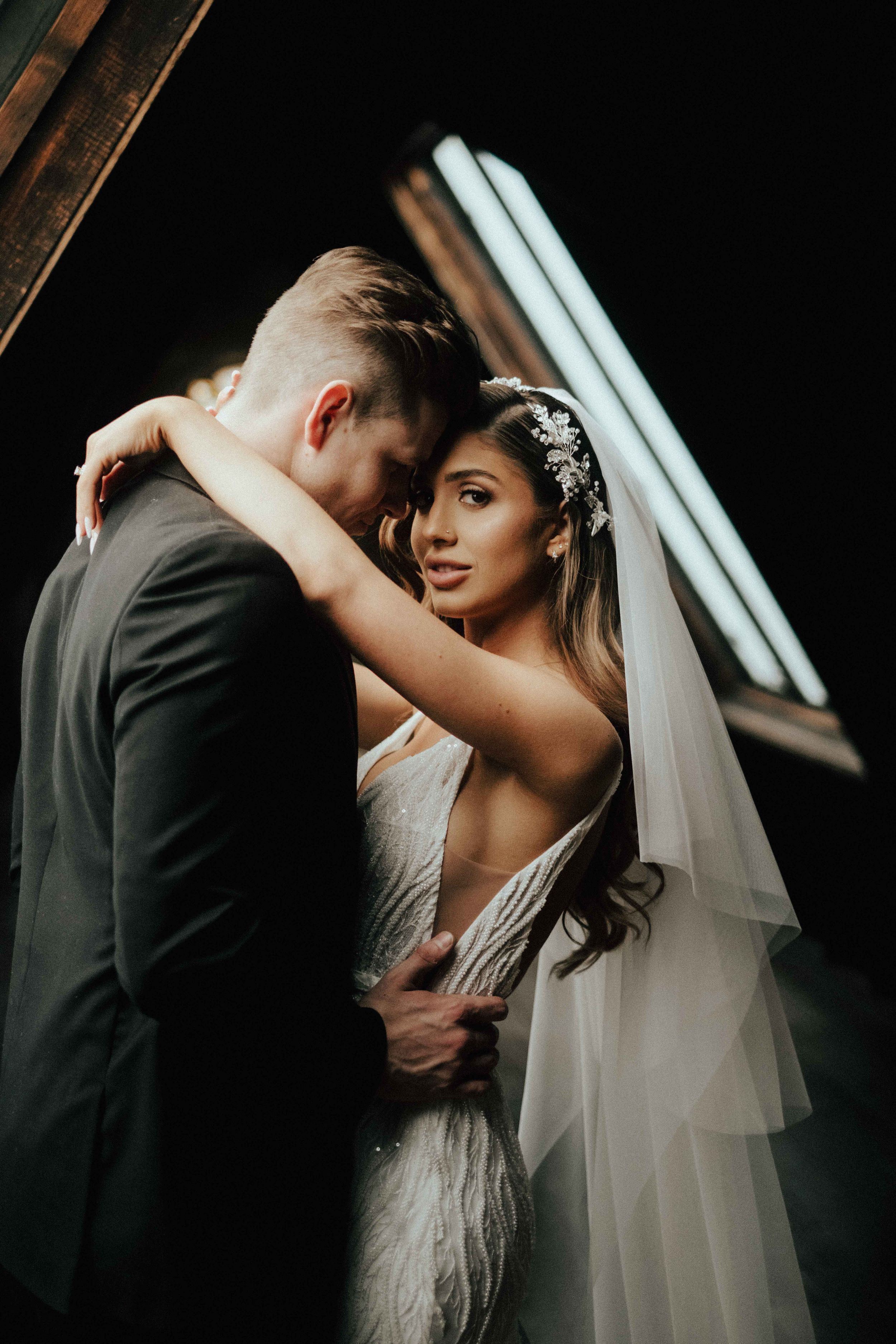 A groom embraces his smiling bride from behind in soft light.
