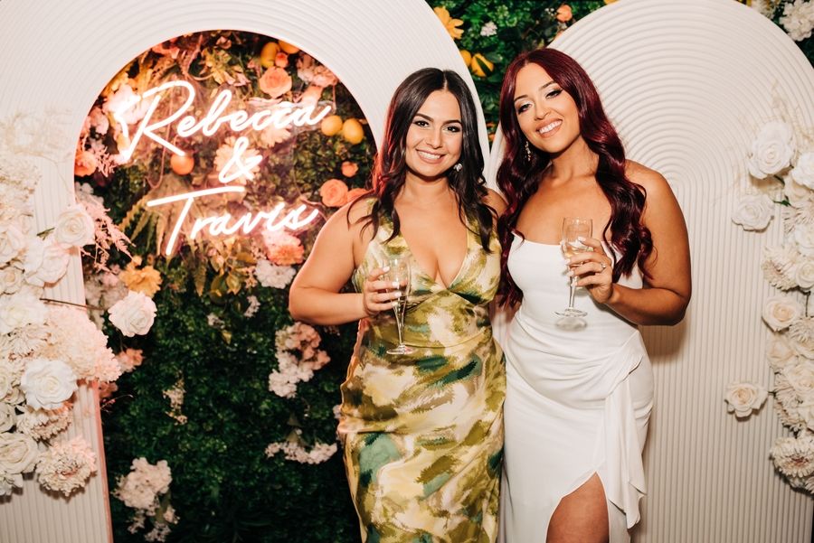 Two people posing by floral arch with drinks at social event.