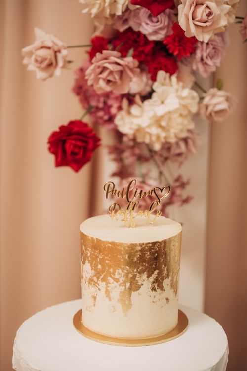 Gold and white celebration cake with floral topper against pink and red rose backdrop.
