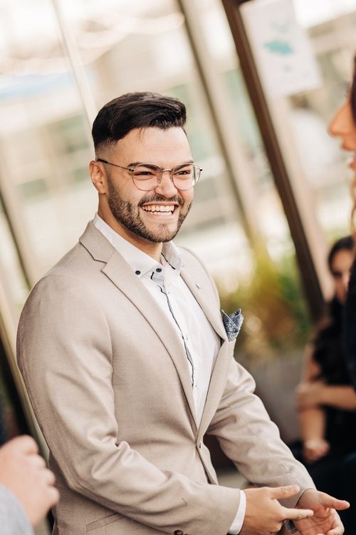 A person wearing glasses and a beige suit jacket smiles warmly at an outdoor event.