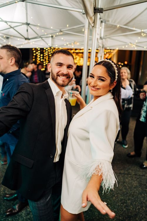 A couple poses together under a tent with string lights at a celebratory event.