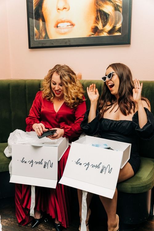 Two friends in formal attire sit on a green couch opening gift bags with excited expressions.