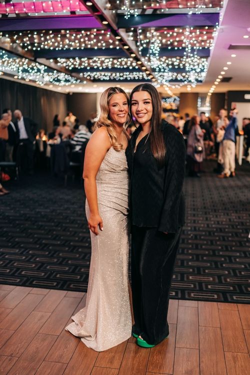 Two friends pose together at a formal event with string lights decorating the ceiling.