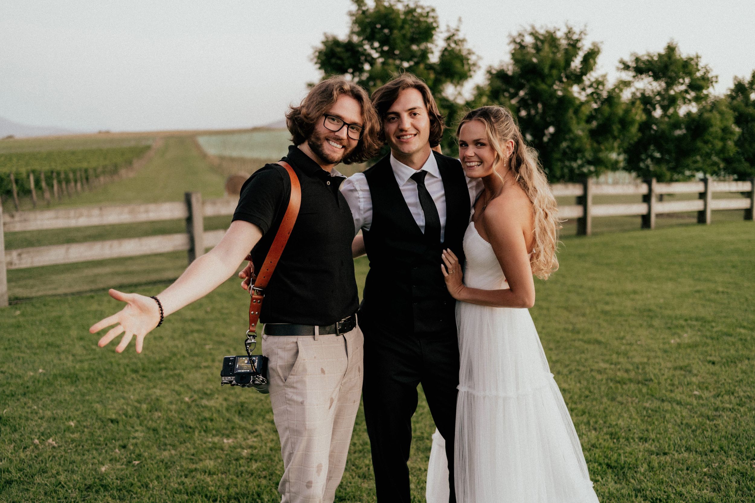 A bride, groom and wedding party smile together in a grassy setting.