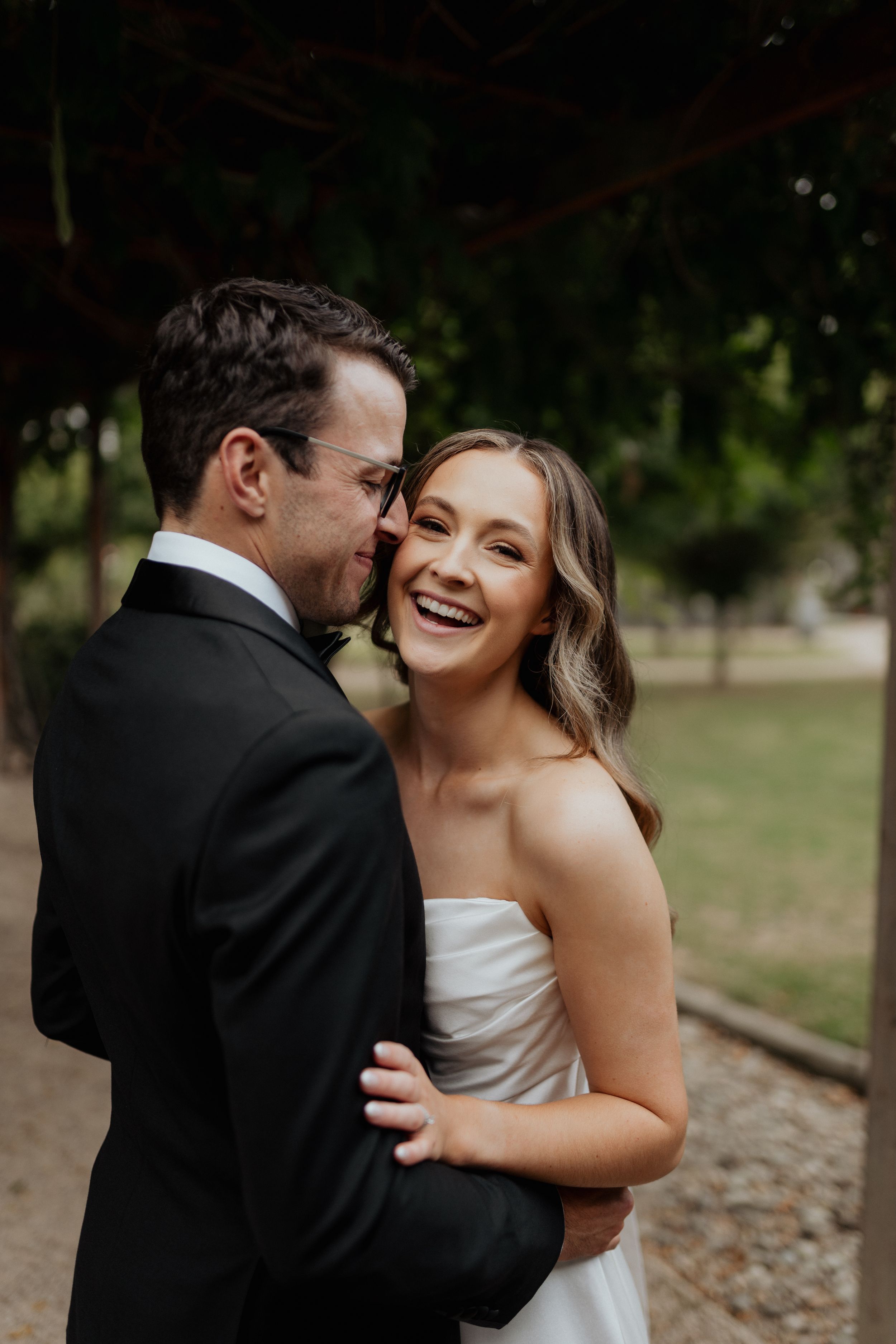 A groom kisses his bride on the cheek in a natural outdoor setting.