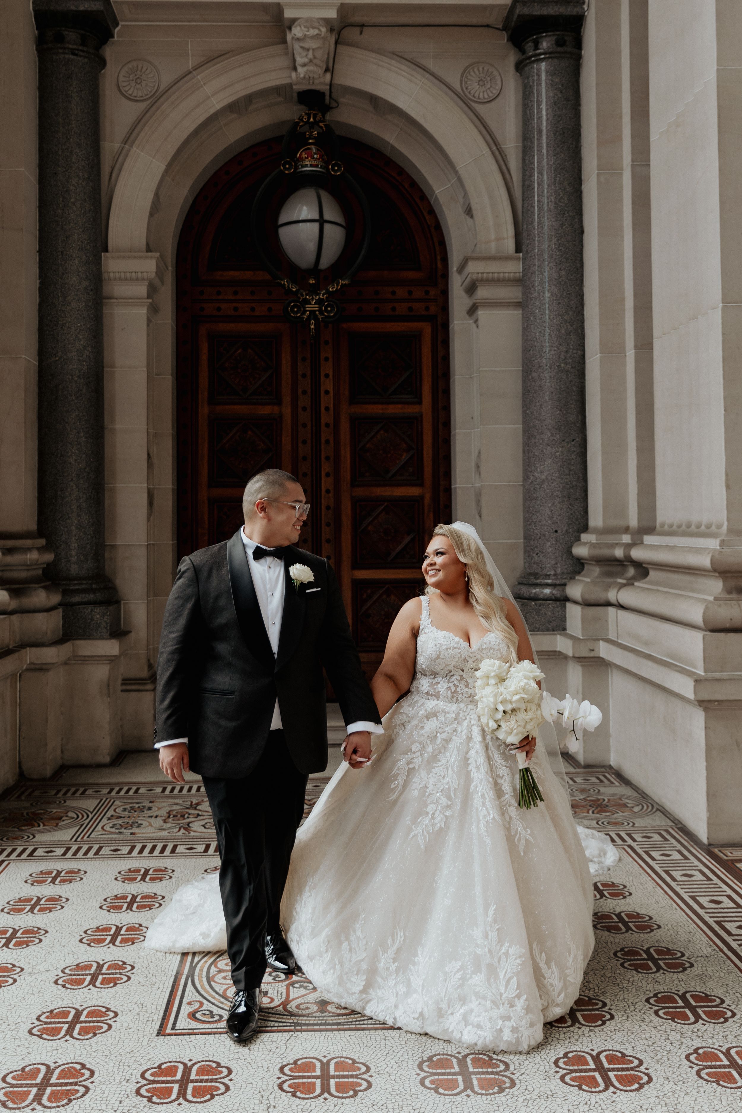 A bride and groom walk hand in hand from a grand stone building.