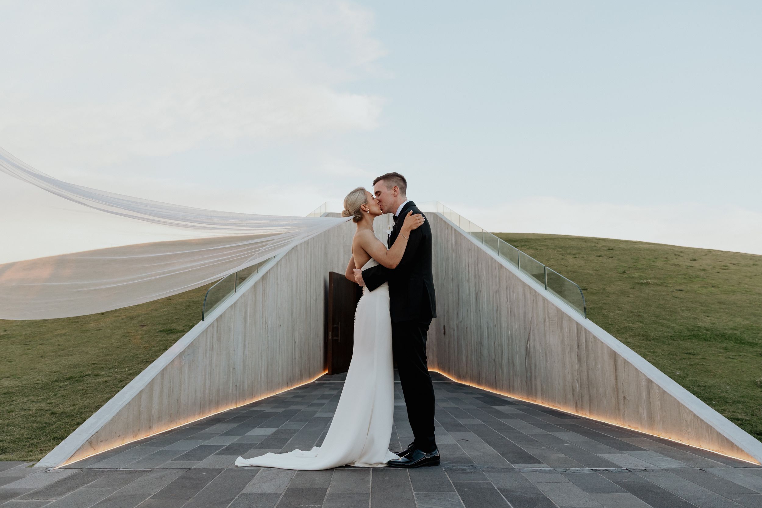 A newly married couple embrace on a modern elevated structure beneath a wide sky.