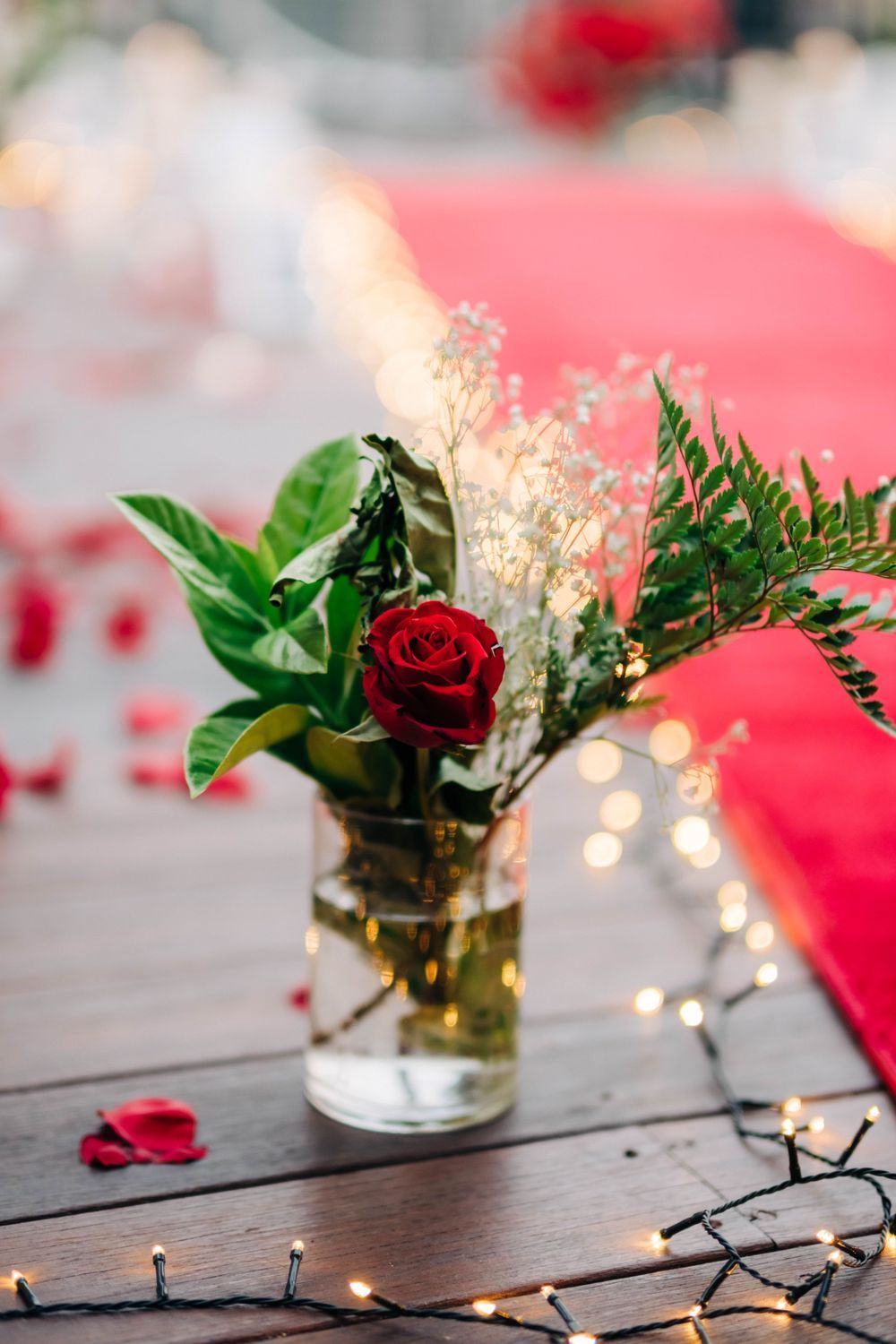 Small flower arrangement with red rose and greenery in glass vase with string lights on wooden surface.