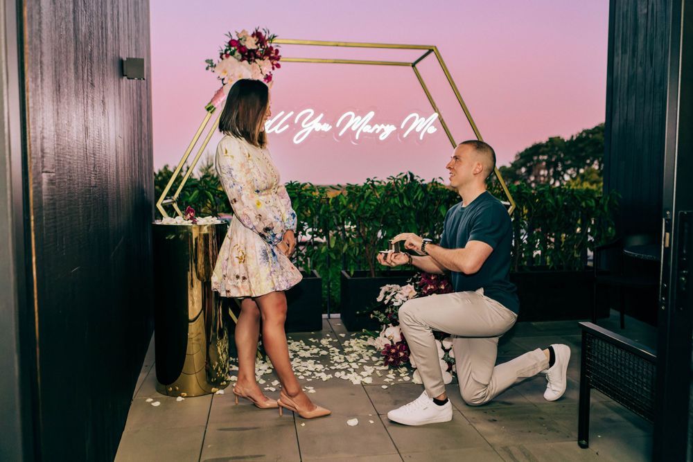 Romantic proposal scene at night with neon sign and string lights.