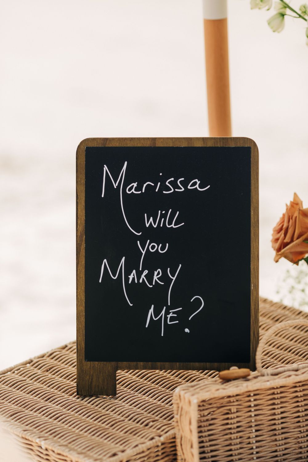 Wooden chalkboard sign with marriage proposal message on wicker basket.