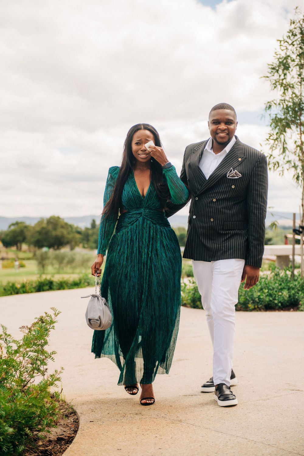 A couple walking outdoors with a woman in a green pleated dress and man in a blazer.