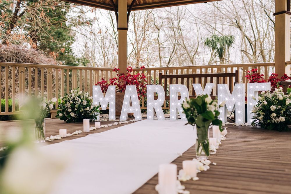 Large white MARRY ME letters displayed on a decorated wooden deck with floral arrangements.