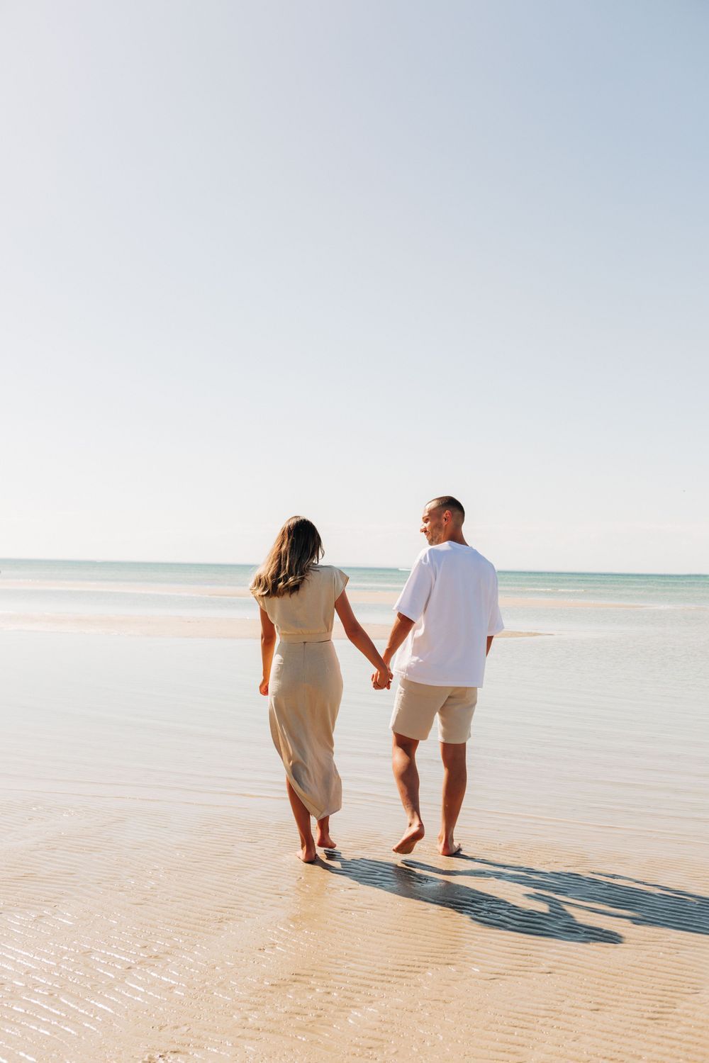 A couple walking hand in hand on a sandy beach during sunset.