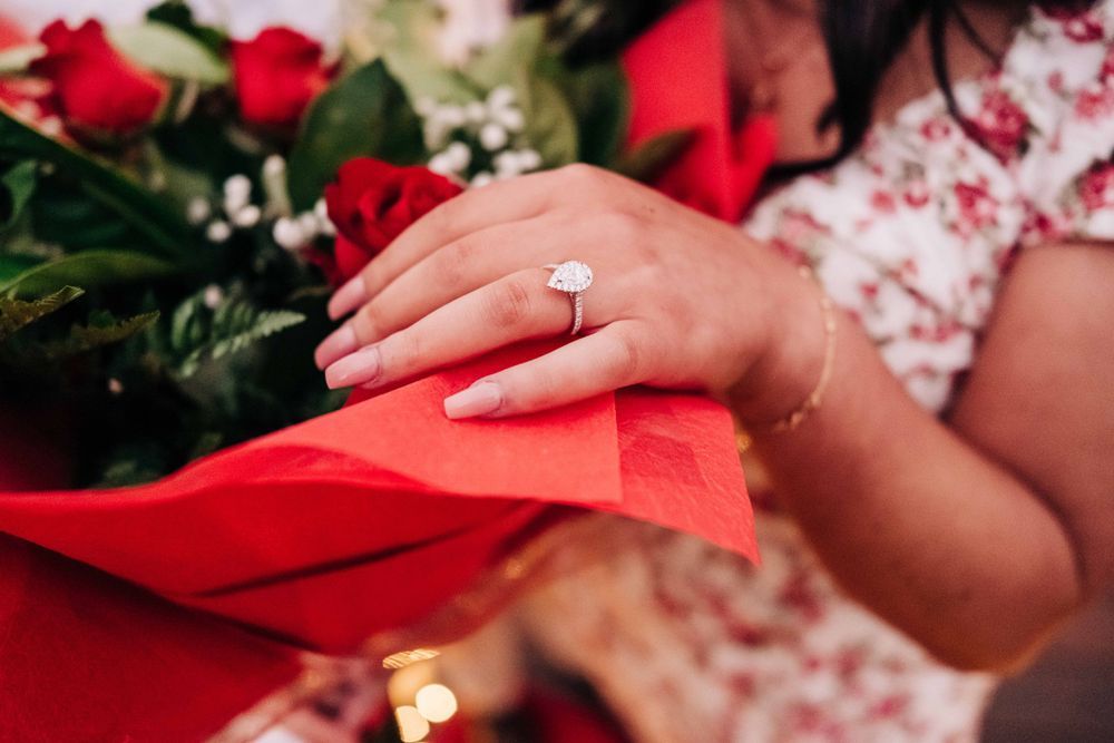 Close-up of a hand wearing an engagement ring with red roses in the background.