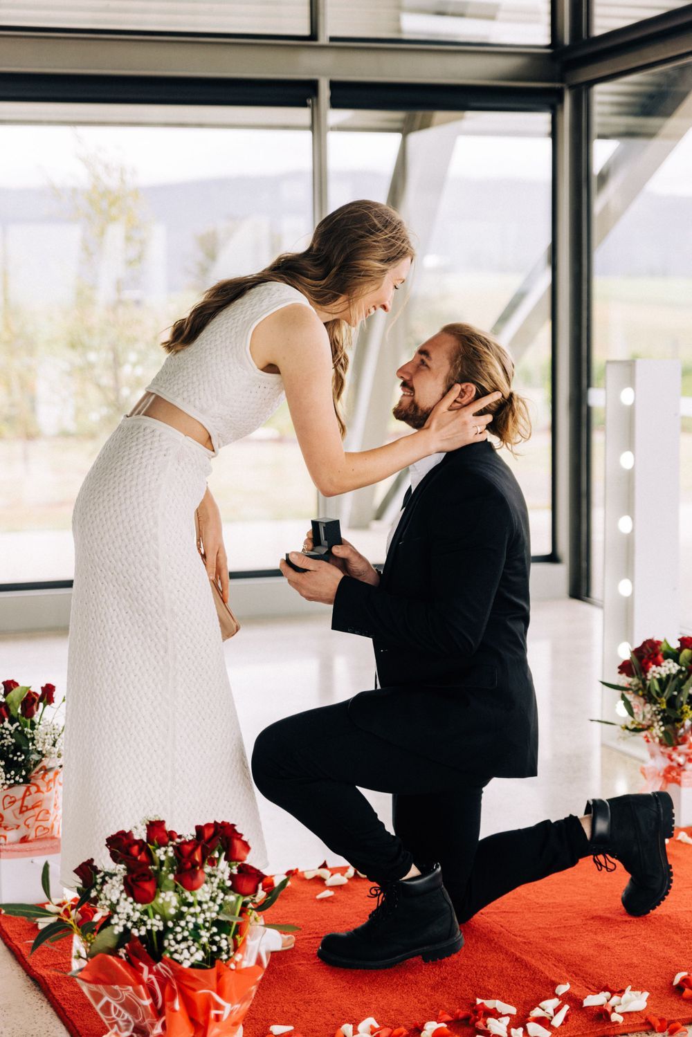 A romantic proposal moment with red rose petals scattered on the floor.