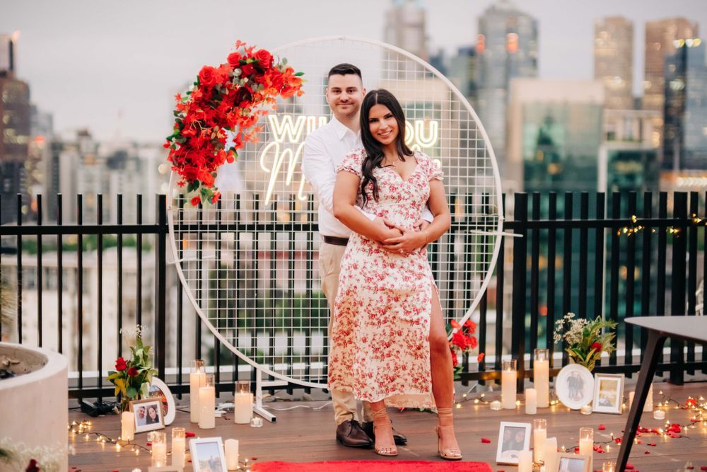 A couple poses by a decorative heart arch with city skyline in background.