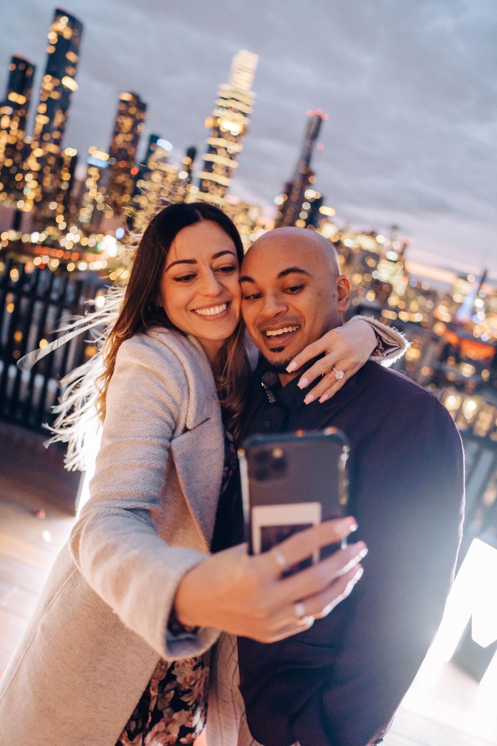 A couple taking a selfie with illuminated city skyline in the background.