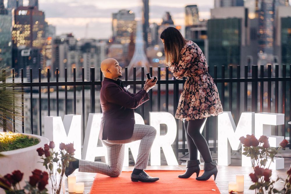 A romantic rooftop proposal with illuminated MARRY ME letters and city skyline.