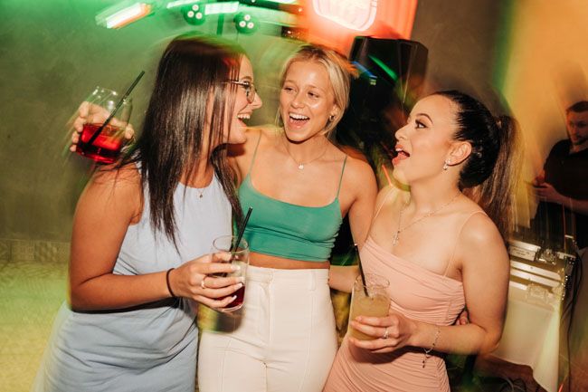 Three friends laughing and holding drinks at a party with colorful lighting.