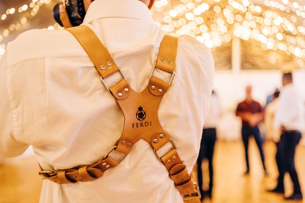 Leather camera strap harness worn on a white shirt at an event with bokeh lights in background.